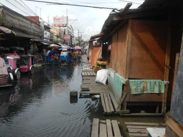 Eine überflutete Stadtstraße mit Fahrzeugen auf der linken Seite, Gebäuden auf beiden Seiten, Strommasten mit Drähten und einem bewölkten Himmel im Hintergrund.