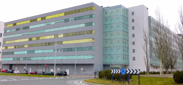 Die Hauptverwaltung der Universität Köln, ein großes Gebäude mit zahlreichen Fenstern, umgeben von Straßenmasten, Straßenlaternen, Kraftfahrzeugen, Schildern, Büschen und Bäumen unter einem klaren blauen Himmel.