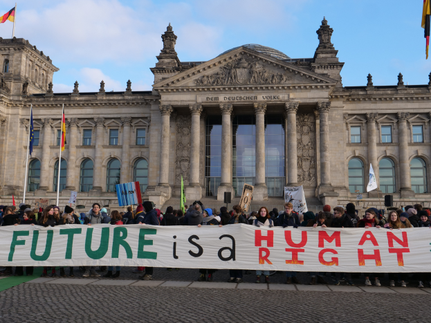 Eine Gruppe von Menschen hält ein Transparent mit der Aufschrift "Zukunft ist ein Menschenrecht" vor dem Reichstaggebäude in Berlin, Deutschland, das mit Fenstern, Säulen und Bögen verziert ist und von Fahnenmasten umgeben ist, unter einem bewölkten Himmel.
