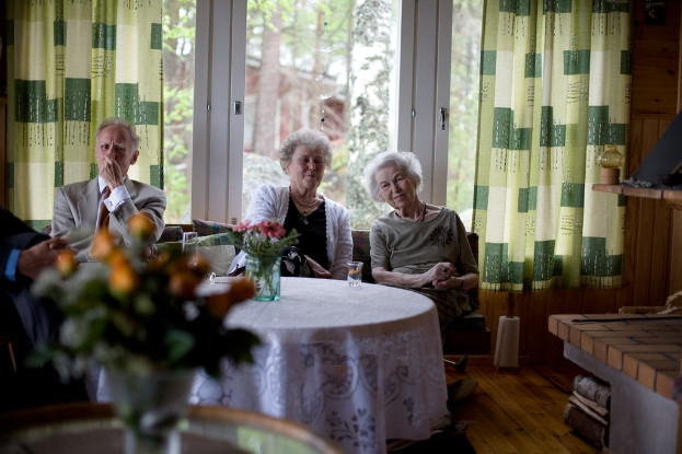 Eine Gruppe älterer Personen sitzt auf einem Sofa mit einem Tisch davor, auf dem eine Blumenvase steht, und einer Glastür mit Vorhängen dahinter.
