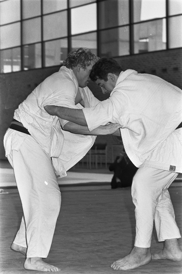 Zwei Männer in weißen Judoanzügen kämpfen intensiv auf einem Boden mit einer Wand und Glasfenstern im Hintergrund, dargestellt in Schwarz-Weiß.