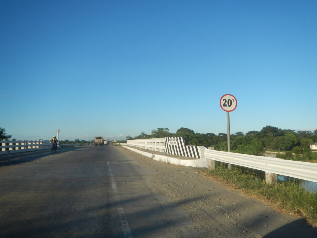 Eine Fahrbahn mit Fahrzeugen, ein Tempolimit-Schild, Geländer, Gras, Bäume und einen klaren blauen Himmel.