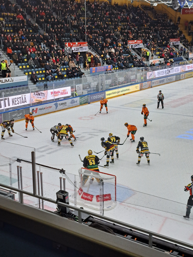 Eine Gruppe von Menschen, die Hockey auf einem Eisring mit einem Netz und einem umliegenden Zaun spielen, Zuschauer sitzen im Hintergrund und Banner mit Text, unter Stadionbeleuchtung.