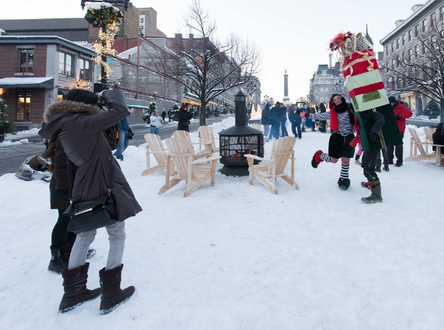 Eine Gruppe von Menschen, die auf Schnee mit Stühlen und Bäumen im Vordergrund, Gebäuden im Hintergrund und dem Himmel darüber steht.