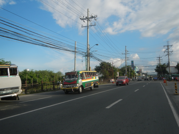 Eine Straßenansicht mit fahrenden Autos und Bussen, umgeben von Strommasten, Laternen, Bäumen und einem bewölkten Himmel.