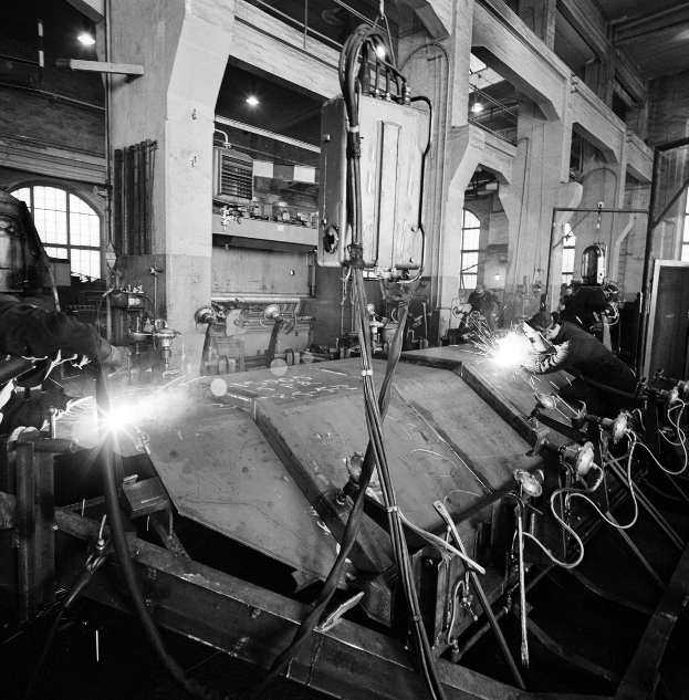 Schwarz-weiß-Foto von Arbeitern, die Metall in einer Fabrik mit Säulen und Fenstern bearbeiten.