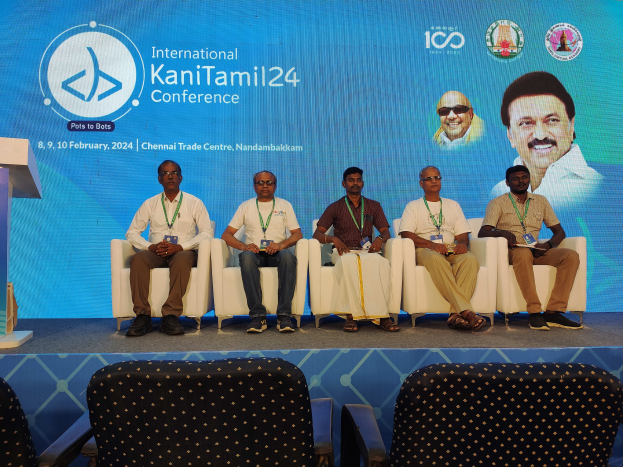 Eine Gruppe von Menschen, die vor einem Podium mit einem Mikrofon sitzen, mit einem Banner im Hintergrund mit Text und Bildern von Menschen und Stühlen unten, das die International Kanitamil24-Konferenz suggeriert.