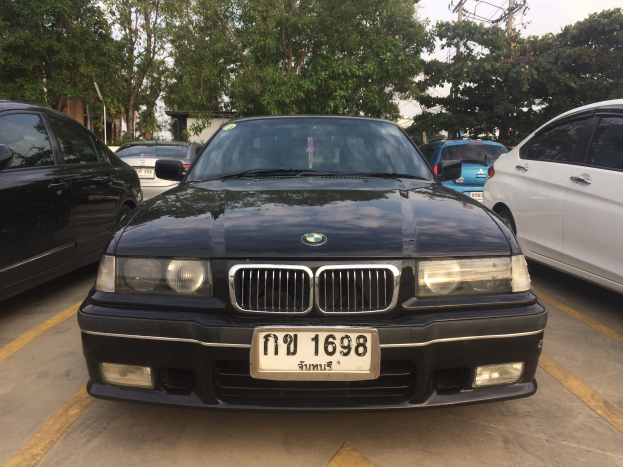 Ein schwarzer BMW E36 318i geparkt auf einem Parkplatz mit anderen Autos, sichtbares Kennzeichen, Bäume, Pfosten, Gebäude und einen klaren blauen Himmel im Hintergrund.