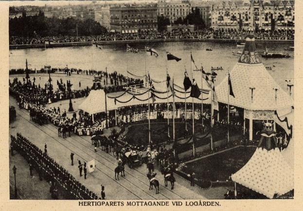 Ein altes Schwarz-Weiß-Foto einer Menschenmenge, die sich um einen Karneval mit Zelten, Fahnen, Stangen und Pferden versammelt hat, mit Gebäuden, Bäumen und Wasser im Hintergrund und einem Text am unteren Bildrand.