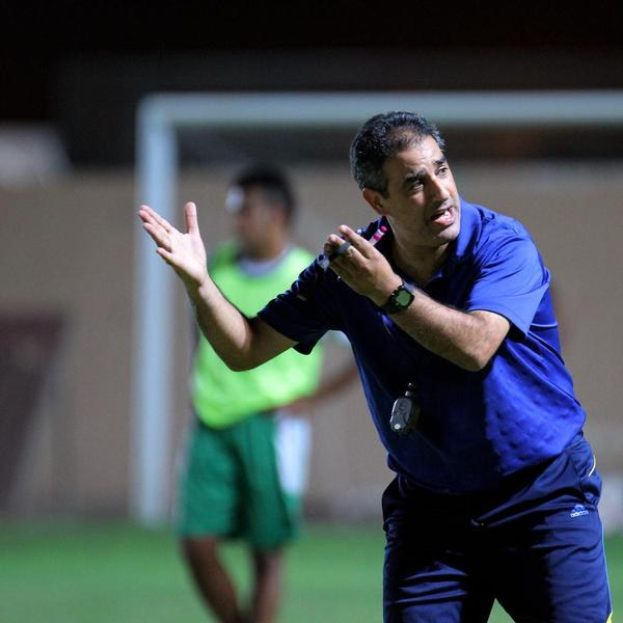 Ein Mann in einem blauen T-Shirt und Hose steht auf einem Fußballfeld und hält ein Mikrofon in der Hand, während er auf Spieler zeigt. Im Hintergrund ist eine Person in einem grünen Kleid und ein Torpfosten zu sehen.