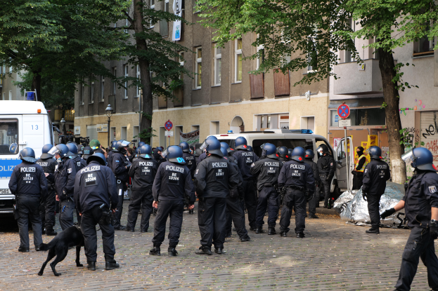 Eine Gruppe von Polizisten in Uniform steht vor einer Menge von Menschen, die Helme tragen und Waffen halten, mit einem Hund im Vordergrund und Fahrzeugen auf der Straße im Hintergrund, eingerahmt von Bäumen und Gebäuden mit Fenstern und Schildern mit Pfosten.