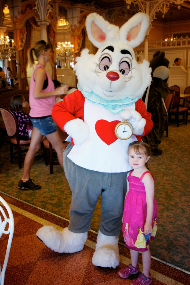 Ein Maskottchen steht neben einem Kind, das eine Uhr hält, mit Menschen im Hintergrund, Stühlen, einem Tisch, einer gestalteten Wand und einem Kronleuchter.