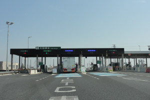 Mautstation an der Straße mit einem vor ihr geparkten Lastwagen, umgeben von weißen und blauen Straßenmarkierungen, Laternenmasten, Schildern und anderen Gegenständen, mit Gebäuden, Bäumen und einem klaren blauen Himmel im Hintergrund.