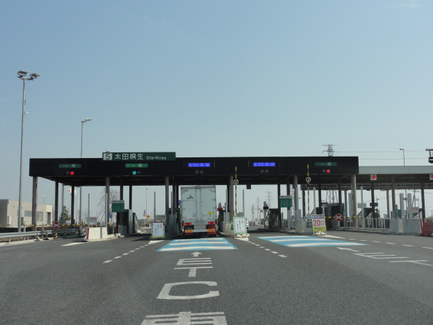 Mautstation an der Straße mit einem vor ihr geparkten Lastwagen, umgeben von weißen und blauen Straßenmarkierungen, Laternenmasten, Schildern und anderen Gegenständen, mit Gebäuden, Bäumen und einem klaren blauen Himmel im Hintergrund.