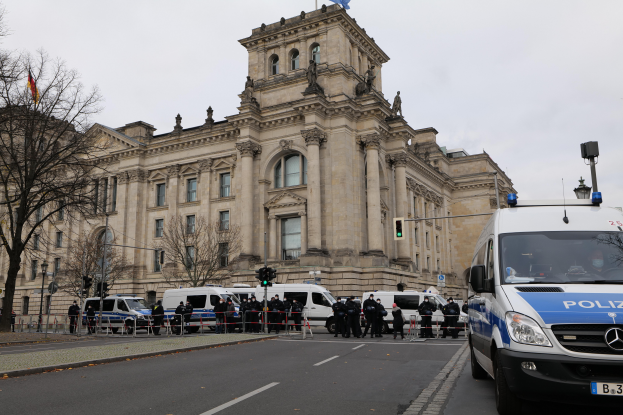 Eine Gruppe von Polizeibeamten steht vor dem Reichstaggeb√§ude in Berlin, Deutschland, mit Fahrzeugen, einem Zaun, Verkehrszeichen, Laternenm√§sten, B√§umen und Flaggen im Hintergrund, unter einem klaren Himmel.