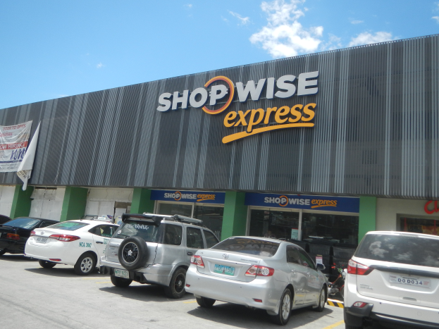 Ein Shopwise Express Geschäft mit parkenden Autos davor, mit einem Banner auf der linken Seite, unter einem bewölkten Himmel.