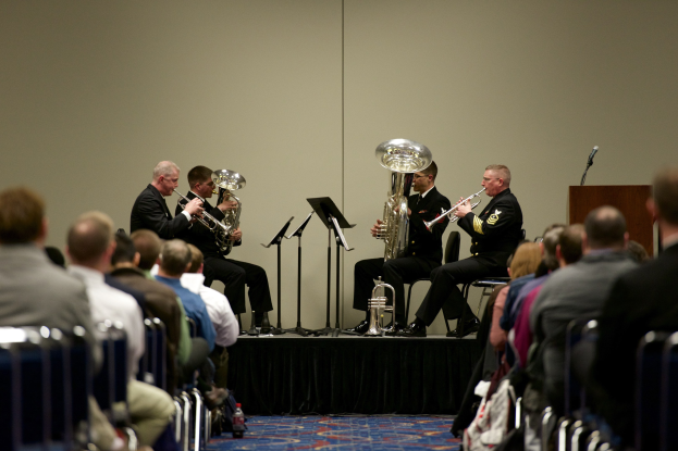 Eine Gruppe von Männern, die auf einer Bühne Brass-Instrumente spielen, mit Notenständen, einem Podium mit Mikrofon und einer sitzenden Zuschauerreihe vor einer Wand.