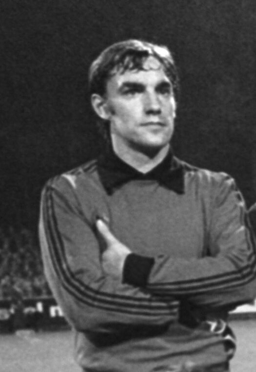 Schwarzes und weißes Foto eines Mannes, der mit verschränkten Armen auf einem Fußballfeld steht, umgeben von sitzenden Zuschauern im Hintergrund.