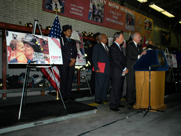 Eine Gruppe von Menschen steht neben einem Podium mit Mikrofonen, Schildern auf Ständern, Fahnen und Schildern an der Wand, alles unter hellen Scheinwerfern.