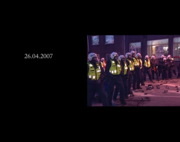 Gruppe von Polizisten in Helmen und Jacken vor einem Gebäude mit Fenstern, mit einem sichtbaren Datum auf der linken Seite.