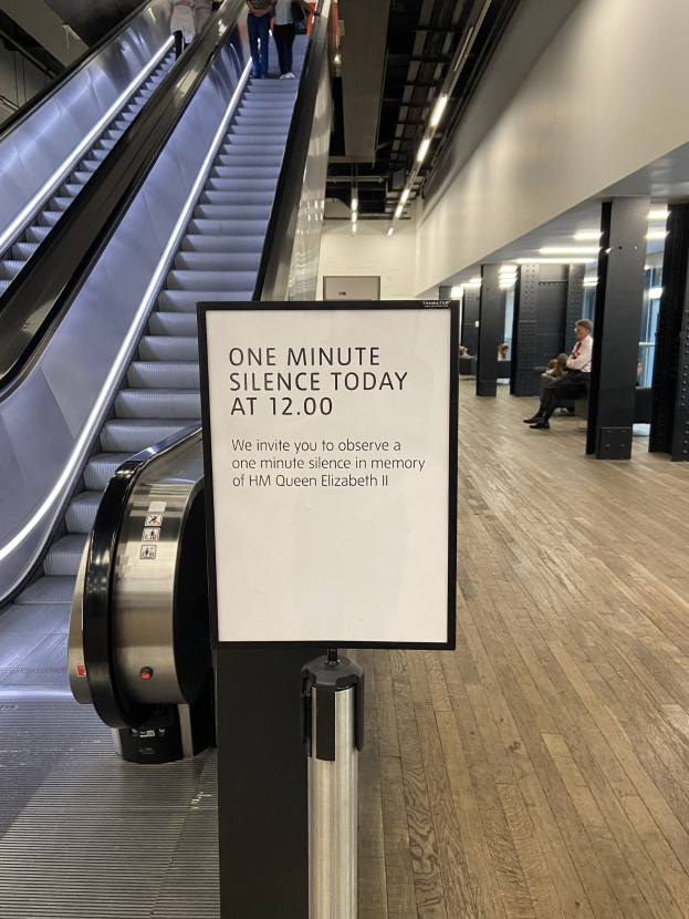 Eine Rolltreppe im Flughafen mit einem Schild, auf dem "Eine Minute Stille heute" steht, sowie einige Personen darauf und Lampen an der Decke im Hintergrund.