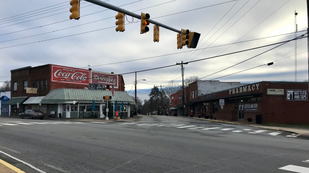 Eine Stadtstraße mit Ampel, Fahrzeugen, Gebäuden mit Namensschildern, Strommasten mit Kabeln, Bäumen und einem bewölkten Himmel.