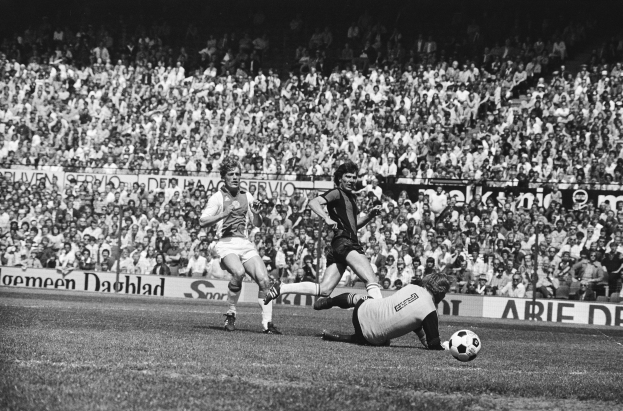 Schwarzes Foto von Männern, die Fussball auf einem Feld mit Zuschauern im Hintergrund und einer Tafel mit der Aufschrift "Bundesliga 1978-79 Ajax 1-1 Hannover 96" spielen.