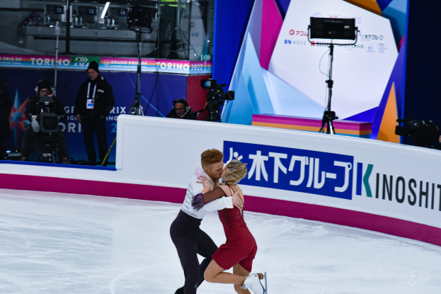 Ein Paar umarmt sich beim Eislaufen auf einer Eisbahn, mit einigen Menschen im Hintergrund, einige halten Kameras, und eine Tafel mit Text, zusammen mit Lichtern und anderen Gegenständen.
