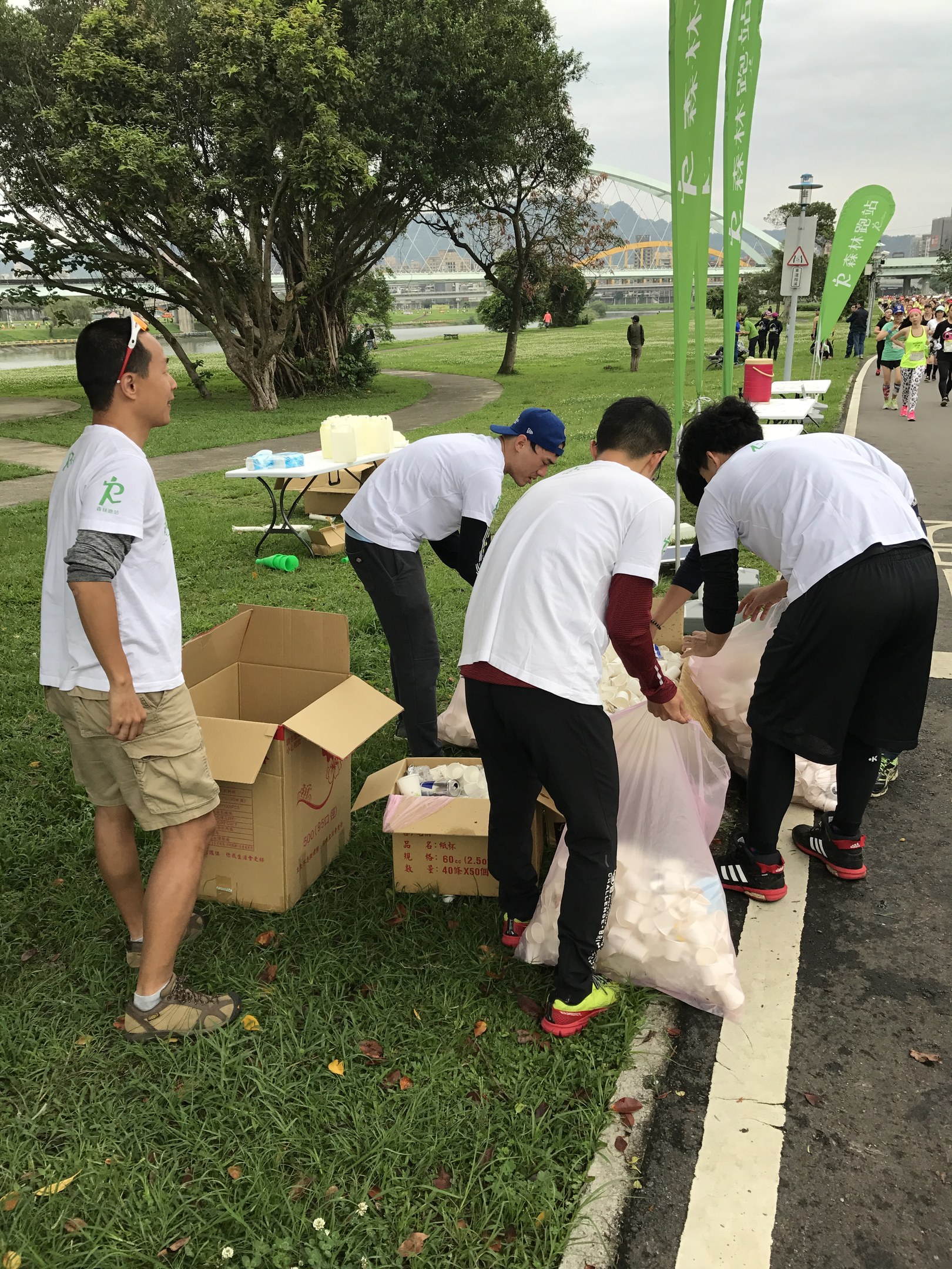 Gruppe von Menschen an einer Straße mit verstreuten Kartonagen und Flaschen, die an einem Spendenlauf teilnehmen mit Bannern, Bäumen, Gebäuden und einem klaren blauen Himmel im Hintergrund.