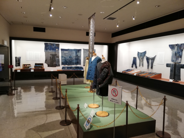 Eine Museumsausstellung mit Puppen in traditioneller japanischer Kleidung, Vitrinen voller verschiedener Kleidungsst├╝cke, Informationsschilder und Deckenleuchten.