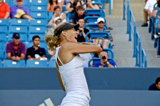 Eine Frau in einem weißen Oberteil und einer schwarzen Mütze steht vor einem blauen Stadionplatz mit Zuschauern.