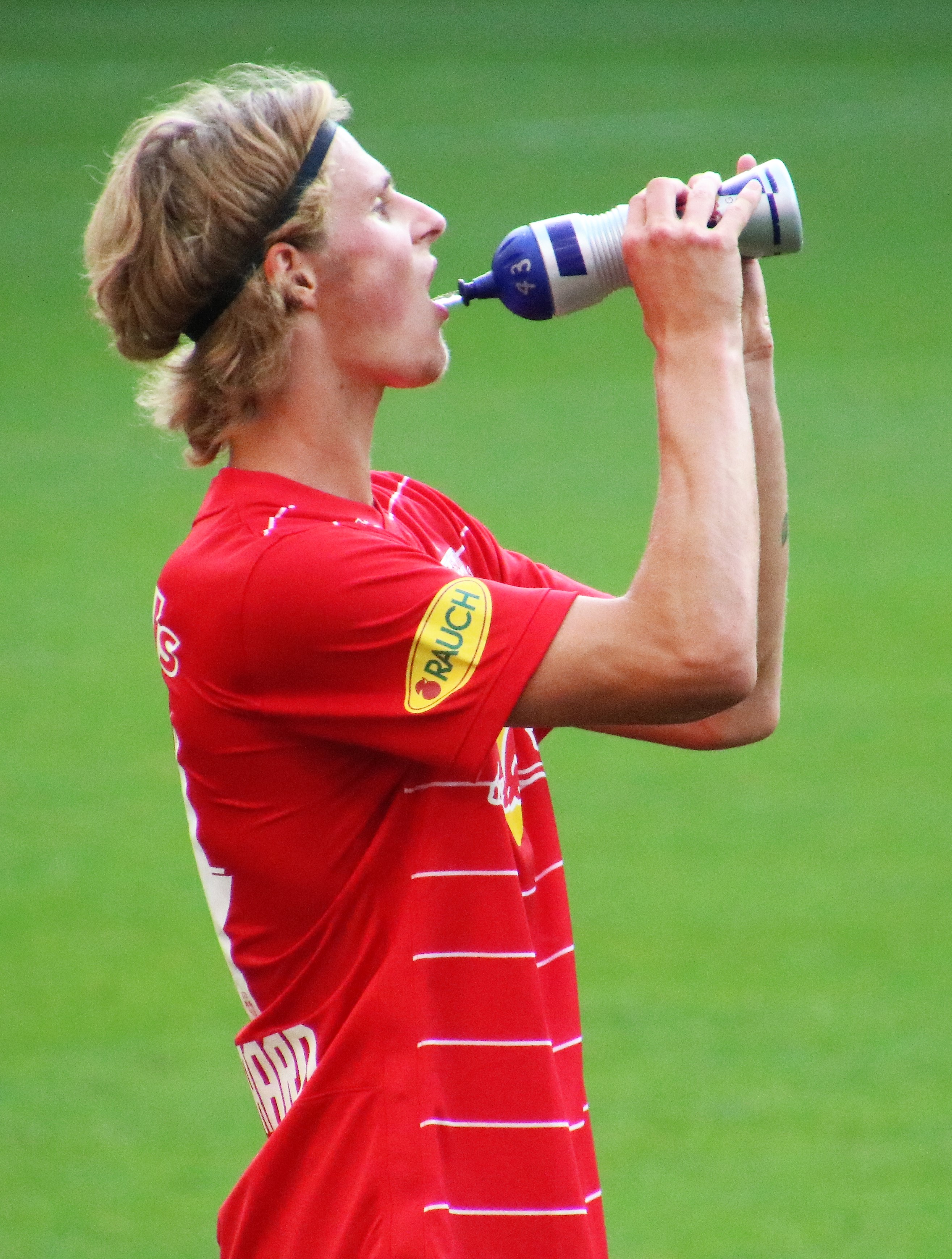 Ein Mann in einem roten T-Shirt trinkt aus einer Flasche, während er auf einem Fußballfeld steht.