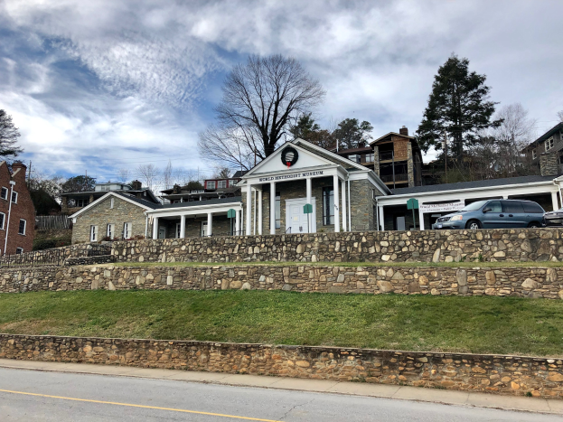Steinwand mit parkenden Autos, umliegende Gebäude mit Fenstern und Türen, Bäume und ein bewölkter Himmel, als Weltkulturerbe-Museum gekennzeichnet.