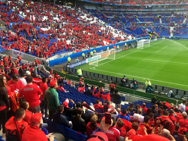 Ein volles Stadion mit Zuschauern, einige sitzen, andere stehen, schauen ein Fußballspiel, mit Bannern, einem Torpfosten und Deckenleuchten.