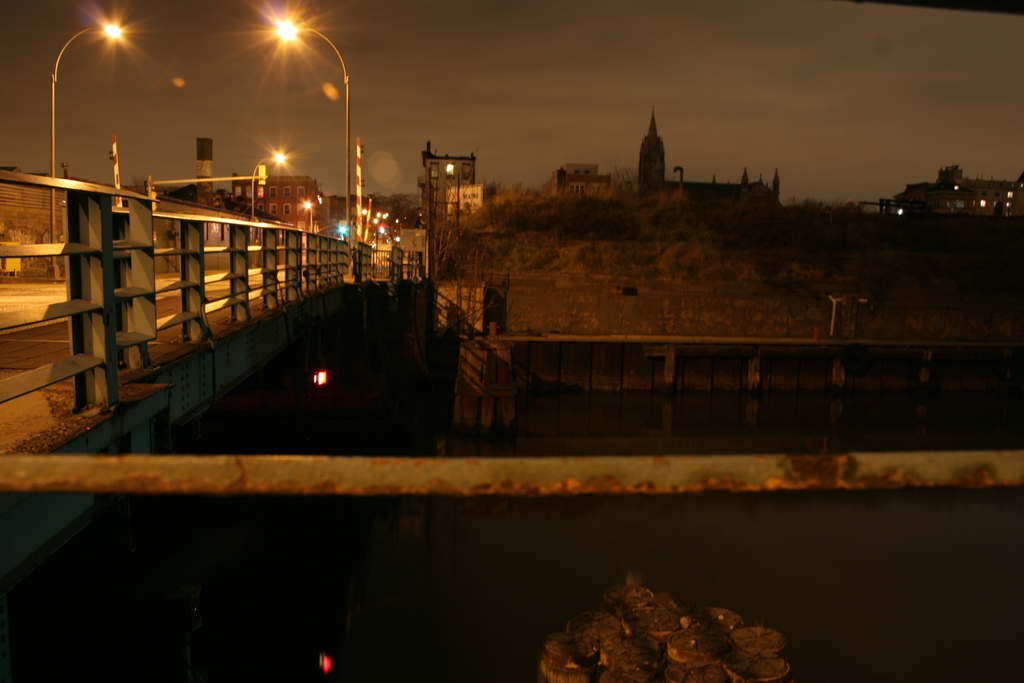 Eine Brücke überspannt einen Fluss mit mehreren Straßenlaternen entlang seiner Länge und zahlreiche Gebäude sind im Hintergrund zu sehen.