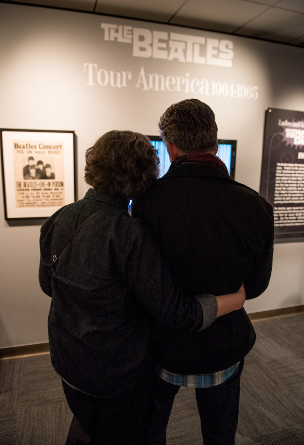 Ein Mann und eine Frau umarmen sich vor einer Wand, auf der ein Fernseher, ein Bilderrahmen und der Text "The Beatles Tour America" zu sehen sind.