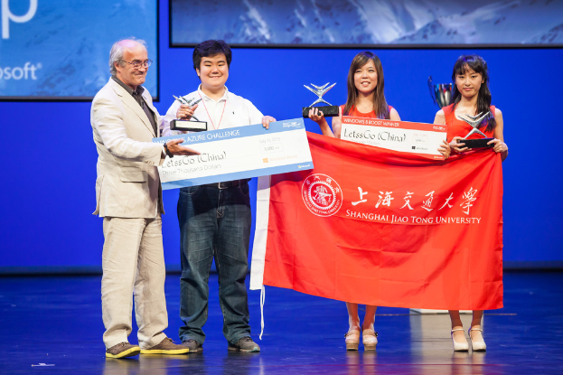 Vier Personen auf einer Bühne mit Auszeichnungen, Schecks und einer Flagge, die lächeln, und ein digitaler Bildschirm mit einer Projektion im Hintergrund.