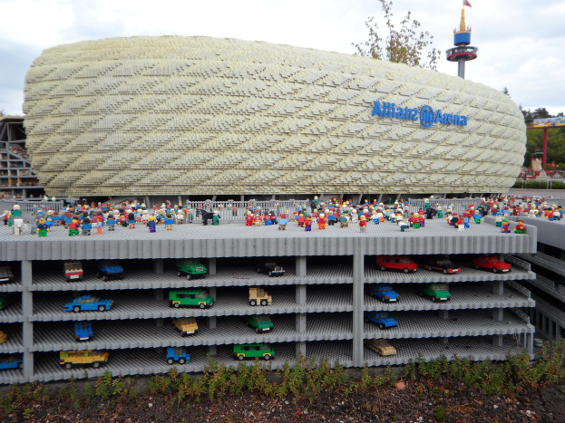 Gro├če Lego-Modell des Allianzstadions in München, Deutschland, mit Menschen davor, umgeben von B├Ąumen, Pfosten, einem Turm und Pflanzen unter einem bew├Âlktem Himmel.