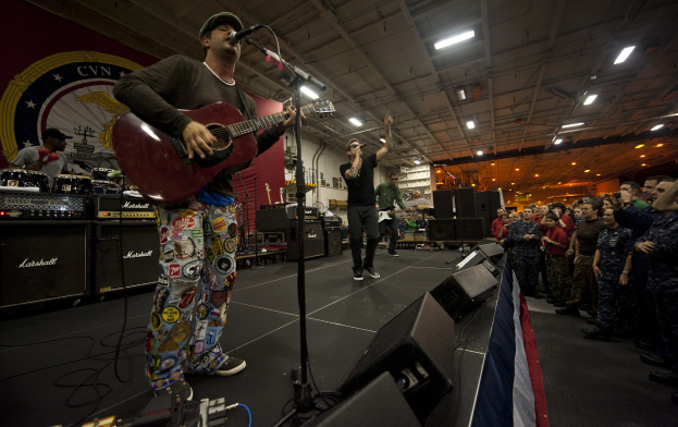 Ein als Chris Stapleton identifizierter Mann spielt eine E-Gitarre auf einer Bühne vor einem Publikum, trägt eine Mütze und ist von einem Mikrofon, Lautsprechern, einer Fahne und anderen Instrumenten umgeben.