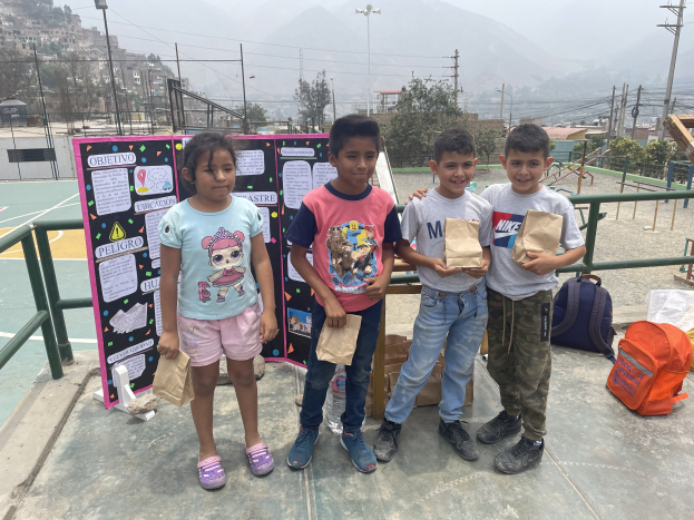 Gruppe von Kindern auf einem Basketballfeld, die Papiertüten halten, mit Vorräten auf dem Boden, einer Tafel mit Text links daneben und einem Zaun, Pfosten und Gebäuden im Hintergrund.