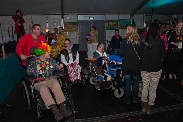 Menschen im Rollstuhl in Kostümen, mit einer Menge im Hintergrund, die verschiedene Gegenstände hält, unter einem Dach mit Lichtern, Rahmen, Brettern, Stangen und einem Tuch.