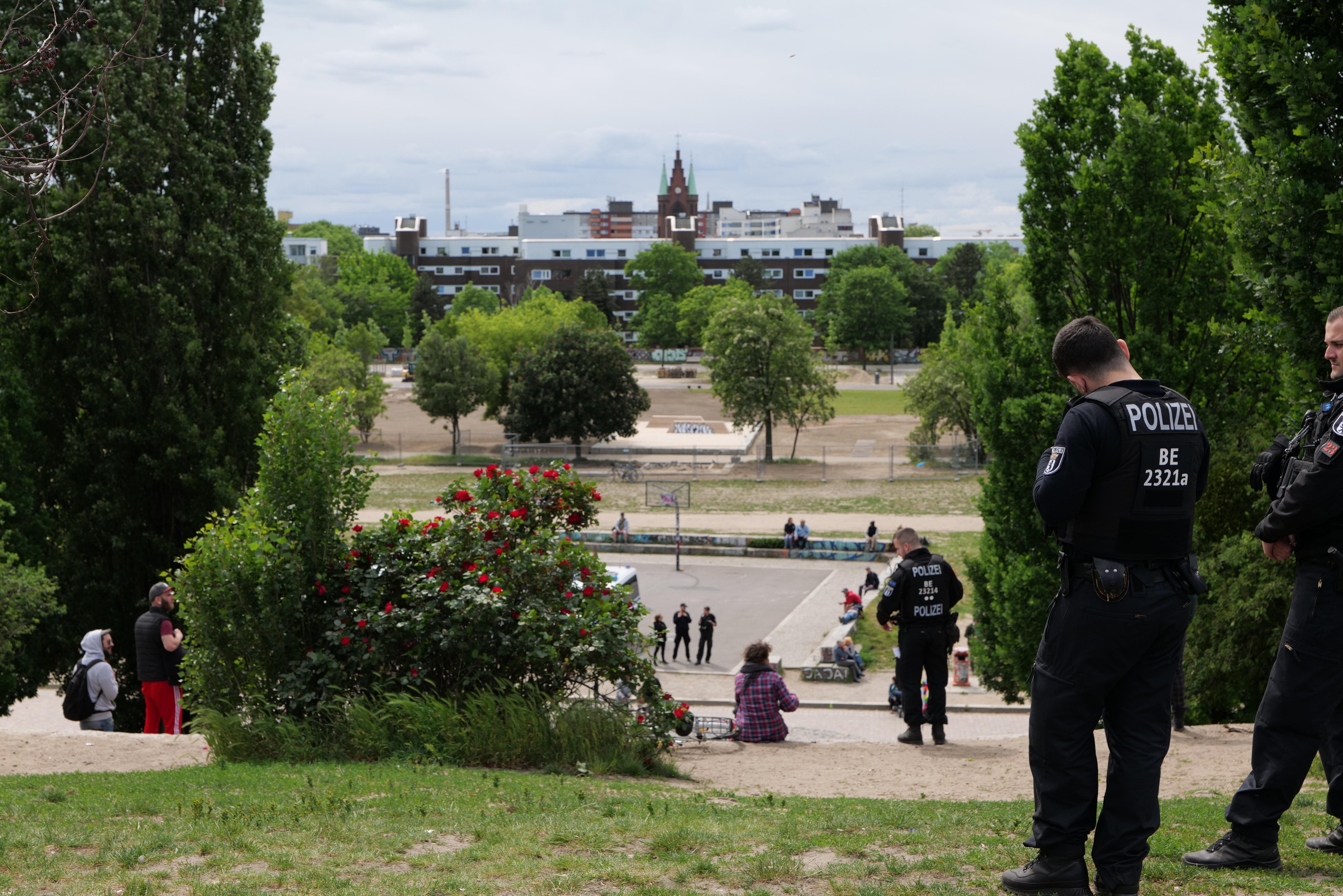 Zwei Polizeibeamte vor einer Gruppe von Menschen in einem Park mit grünem Gras, Bäumen, bunten Blumen, Gebäuden, Polen und einem klaren blauen Himmel.