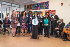 Gruppe von Menschen vor einem Podium mit Mikrofon und einer Tafel mit dem Text "Sancho Stadtverordneter tritt dem Stadtrat bei", mit sitzenden und stehenden Gästen, Flaggen, Feuerlöscher und Deckenbeleuchtung im Hintergrund.