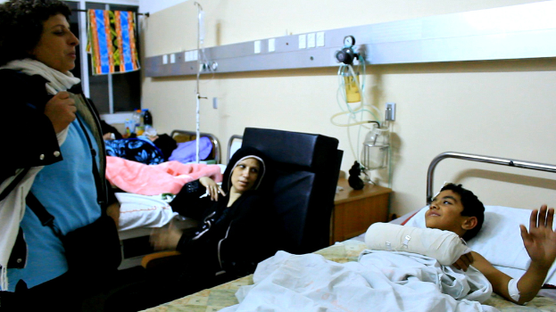 Ein Krankenhauszimmer mit mehreren Betten, die von Patienten belegt sind, Infusionsflaschen auf Ständern zwischen den Betten und eine Frau mit einer Tasche in der Nähe eines der Patienten.