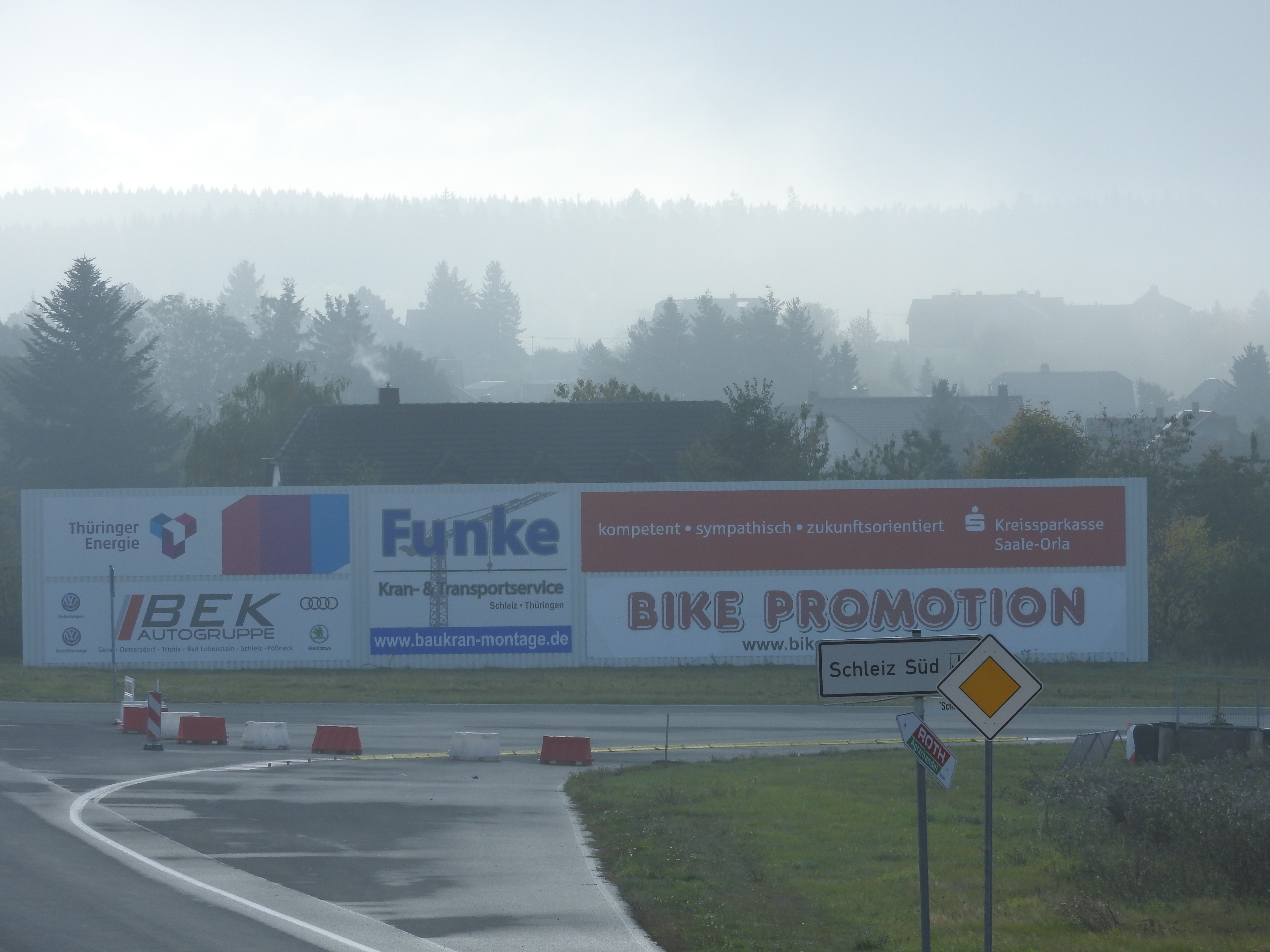 Eine Straße mit einem "Bike Promotion"-Schild auf der linken Seite, Gras und Pflanzen auf der rechten Seite und Bäumen, Gebäuden und einer Werbetafel im Hintergrund unter einem sichtbaren Himmel.
