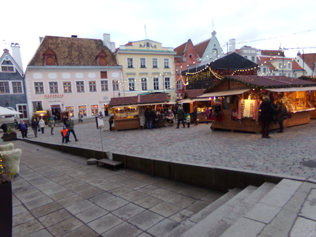 Ein lebendiger Weihnachtsmarkt in Tallinn, Estland, mit Menschen um geschmückte Stände, festlicher Beleuchtung und Gebäuden mit Fenstern im Hintergrund, unter einem bewölkten Himmel, mit Stufen, die zum Markt führen und Töpfen mit Pflanzen, die herumstehen.