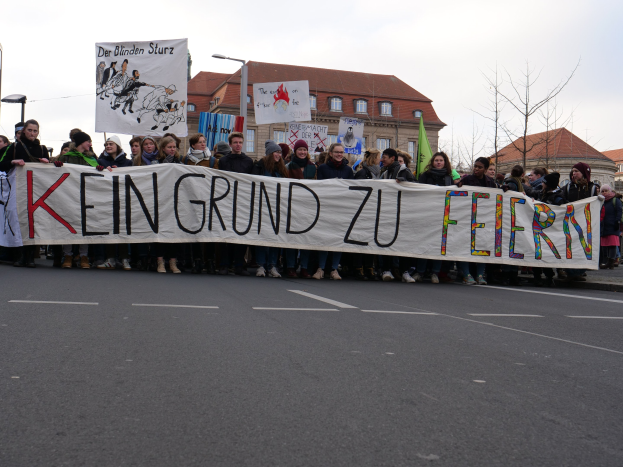 Demonstranten mit einem Banner "Kein Grund zu Feiern" gegen deutsche Sparmaßnahmen, mit Straßeninfrastruktur und Gebäuden im Hintergrund unter einem klaren Himmel.