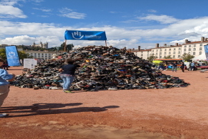 Eine Gruppe von Menschen steht um einen Haufen Schuhe auf einem Feld mit Gebäuden, Bäumen und einem bewölkten Himmel im Hintergrund bei einem internationalen Schuh-Recycling-Event.