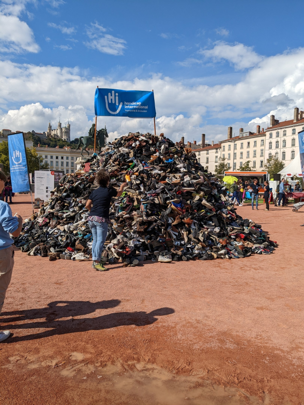 Eine Gruppe von Menschen steht um einen Haufen Schuhe auf einem Feld mit Gebäuden, Bäumen und einem bewölkten Himmel im Hintergrund bei einem internationalen Schuh-Recycling-Event.