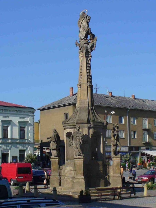 Ein Denkmal steht auf einem Platz in einer Stadt umgeben von Gebäuden, Straßenlaternen, Fahrzeugen, Fußgängern, Bänken, Pflanzen und einem bewölkten Himmel.